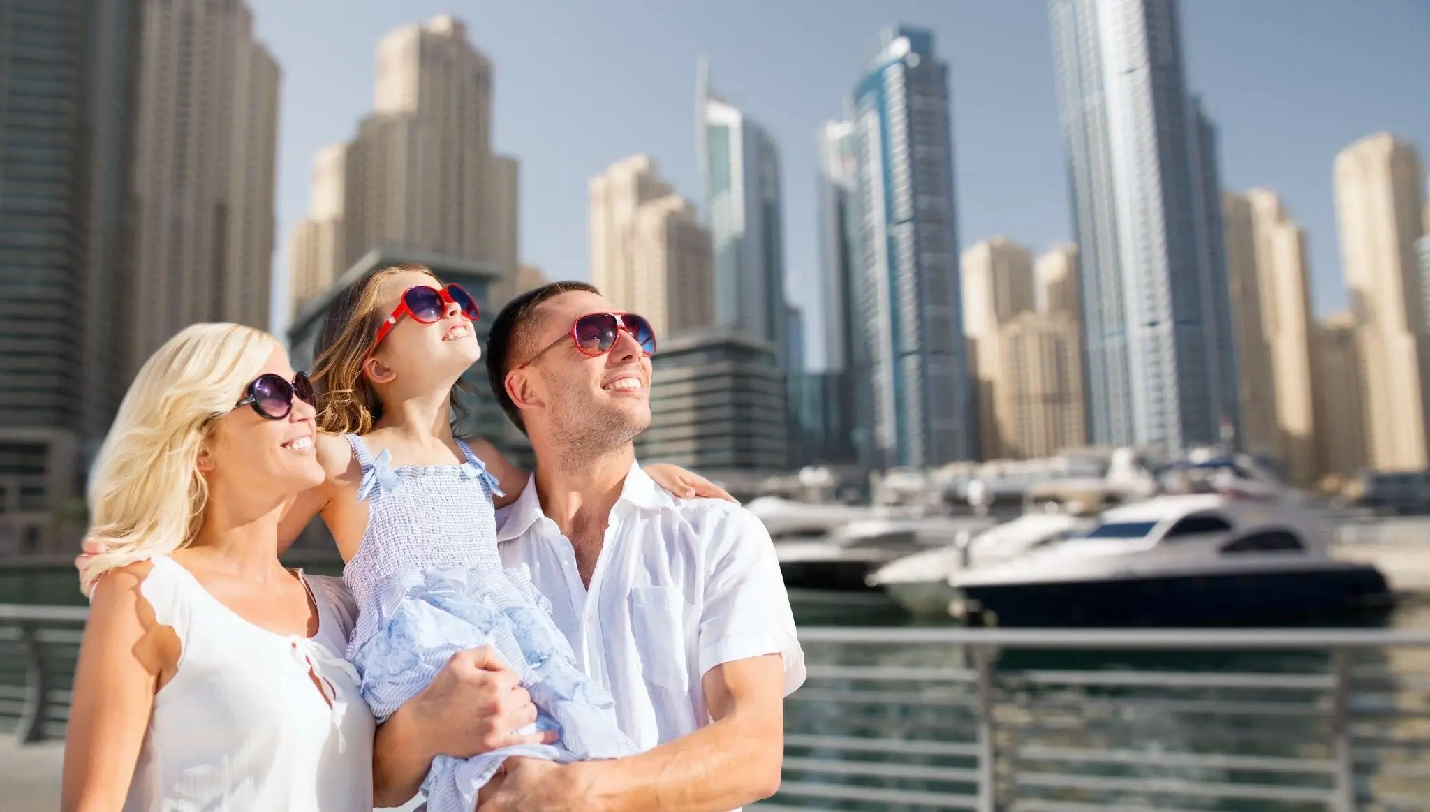 A family stands together in front of a marina, with a yacht visible in the background.