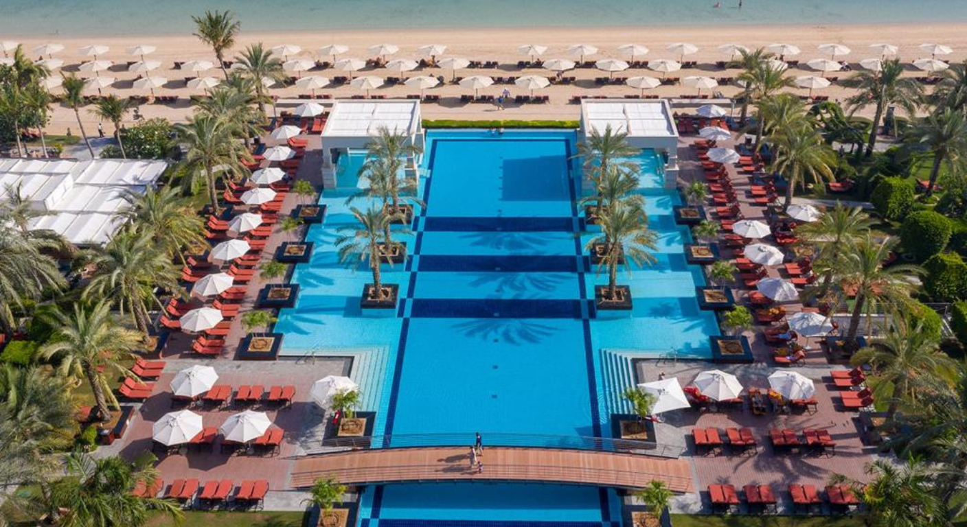 Aerial view of Jumeirah Zabeel Saray Dubai featuring a large beachfront swimming pool, palm-lined decks, and private beach loungers