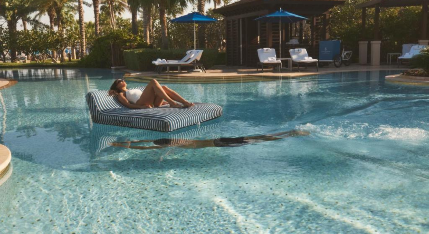 Guest relaxing on a floating daybed in the outdoor swimming pool at Four Seasons Resort Dubai at Jumeirah Beach, surrounded by palm trees