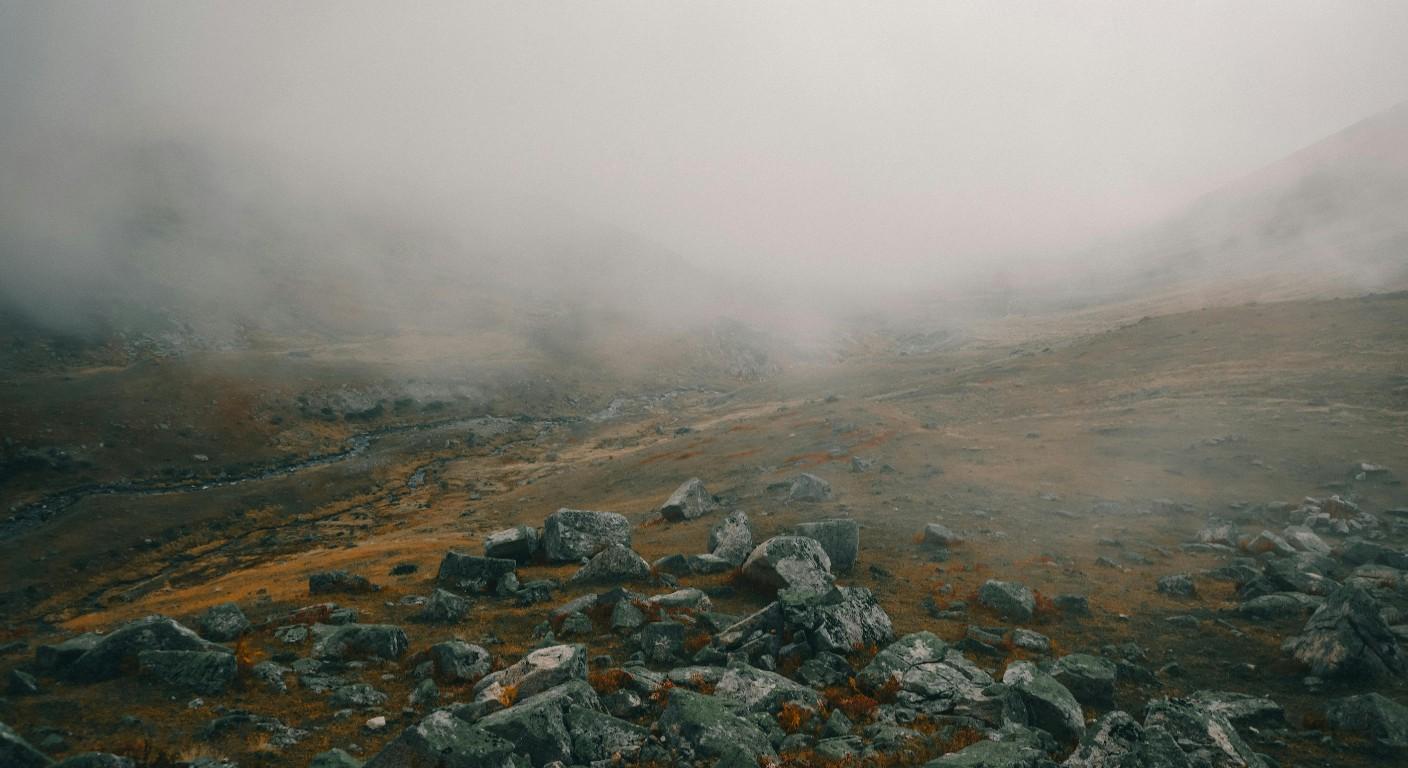 A foggy mountain valley with scattered rocks and muted brown grass.