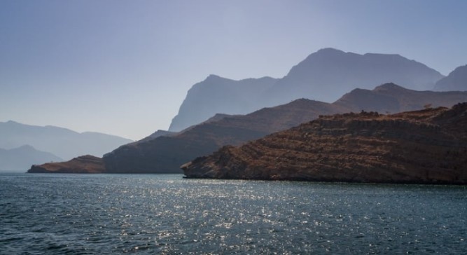Khasab Tours: Panoramic view of a peaceful turquoise bay in Musandam with a small fishing boat and rugged limestone cliffs at golden hour.
