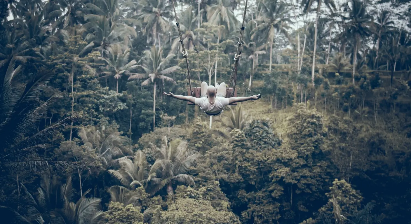 A person swinging high above a lush green jungle with tall palm trees in the background, arms stretched wide in mid air.