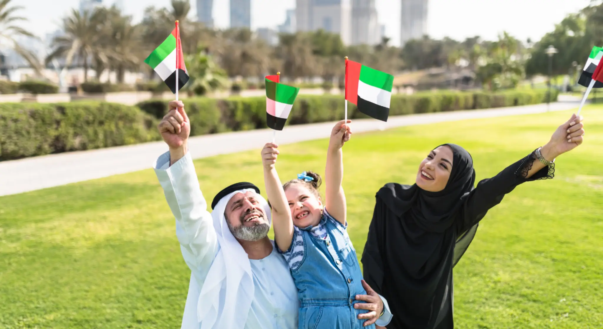Happy family waving UAE flags in a park with city skyline in the background/