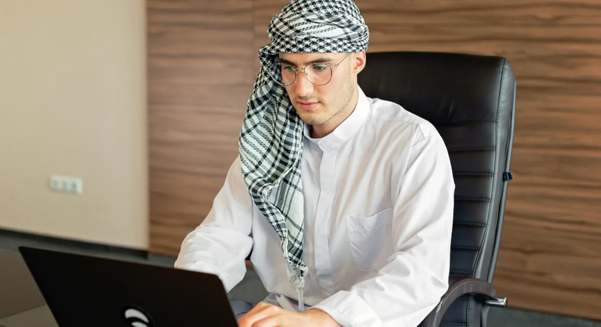 Arab man in traditional attire working on laptop at office desk in UAE