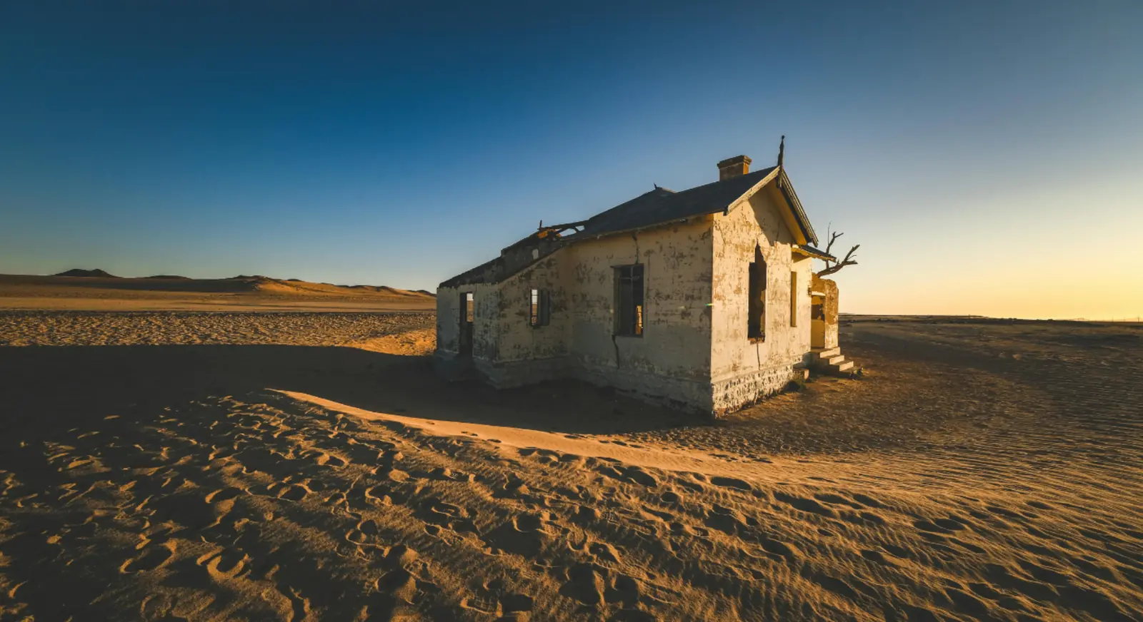 Abandoned house surrounded by desert sand at sunset