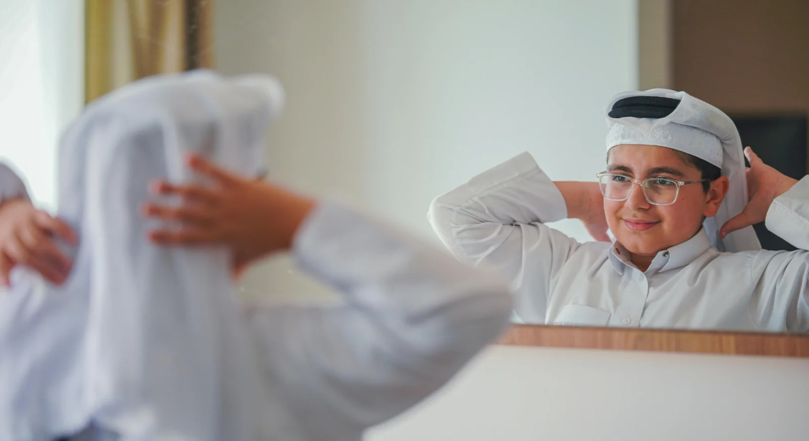Young Emirati boy wearing traditional kandura and ghutra in front of mirror