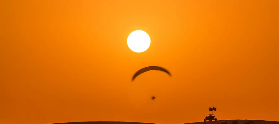 Paraglider above a desert camp at sunset, with tents and vehicles on the horizon.