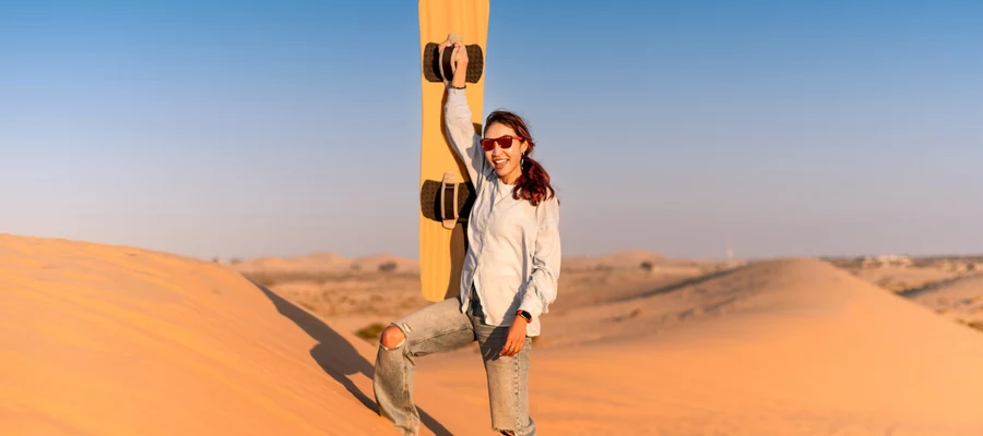 Woman carrying a sandboard on a sunlit desert dune in Dubai.